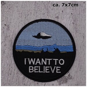 Könnte beinhalten: Ein gestickter Patch in Schwarzweiß mit einem UFO, das über einer Landschaft fliegt. Der Text "I WANT TO BELIEVE" steht unter dem UFO. Der Patch hat einen Durchmesser von etwa 7 cm.