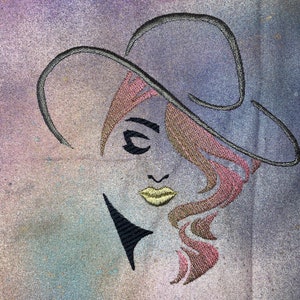 May include: Embroidered design of a woman's face with pink and brown hair, wearing a black hat. The background is a light purple and blue watercolour wash.