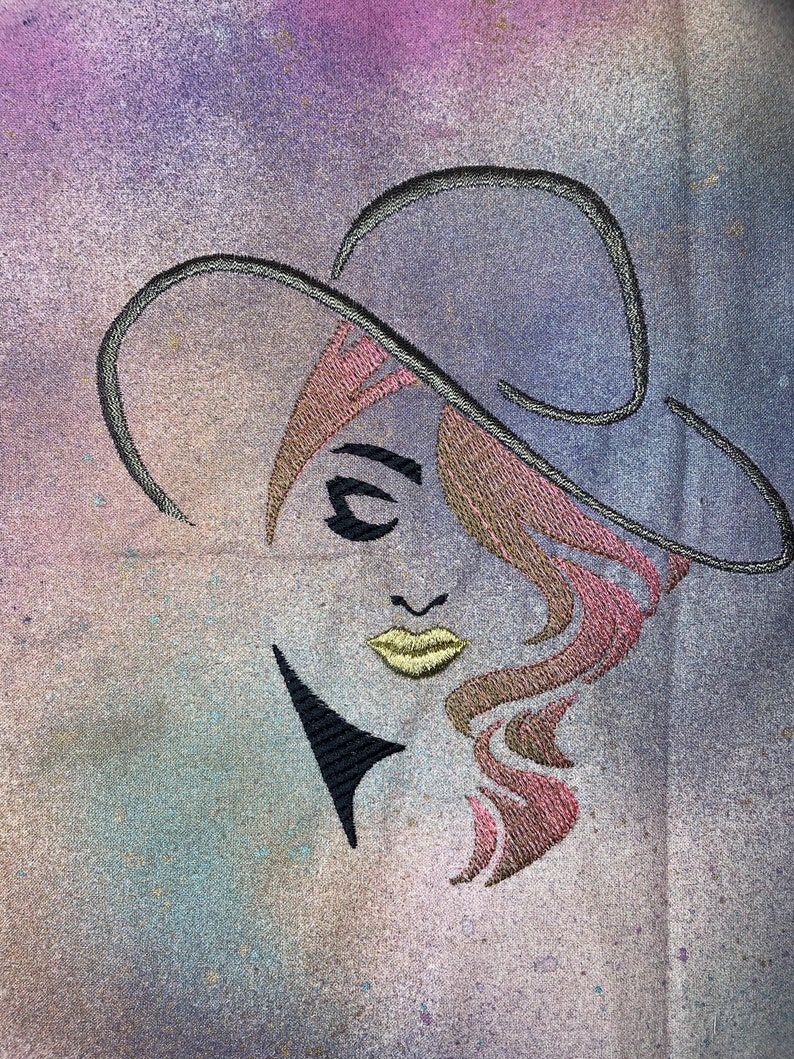 May include: Embroidered design of a woman's face with pink and brown hair, wearing a black hat. The background is a light purple and blue watercolour wash.