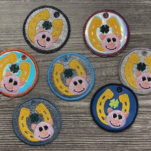 Könnte beinhalten: Acht runde, glitzernde Filzpatches mit einem goldenen Hufeisen, einem Kleeblatt und einem rosafarbenen Schweinegesicht. Die Patches sind in verschiedenen Farben, darunter Silber, Blau und Rot.
