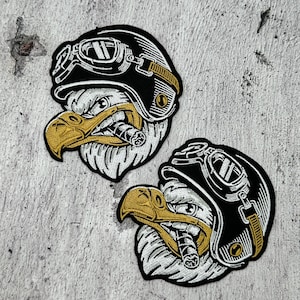 Könnte beinhalten: Zwei gestickte Aufnäher mit einem Adlerkopf, der einen schwarzen Fliegerhelm mit Brille trägt. Der Adler hat einen goldenen Schnabel und raucht eine Zigarre. Die Aufnäher sind weiß, schwarz und gold.
