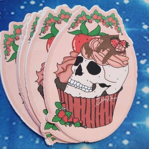 May include: A pink cupcake with a skull and pink frosting topped with strawberries and chocolate. The cupcake is surrounded by a wreath of strawberries and leaves.