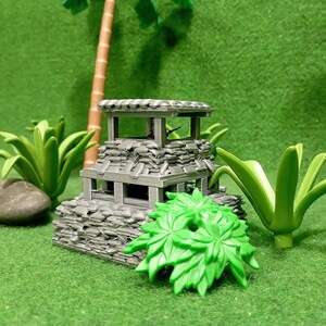 以下が含まれることがあります： 緑色のプラスチック製の植物に囲まれた、わらぶき屋根の灰色の石造りの見張り塔。見張り塔は、卓上ゲームやジオラマ用のミニチュアモデルです。