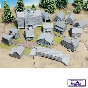 Può includere: Una collezione di edifici in miniatura grigi, tra cui case, fienili e una struttura più grande con più finestre e una cupola centrale. I modelli sono disposti su una superficie beige con alberi in miniatura.