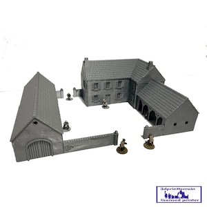 Op de afbeelding: Grijs model van een Europees dorp, bestaande uit een huis, een schuur en muren. De gebouwen hebben gedetailleerde dakpannen en luiken. Kleine beeldjes zijn rond de structuren geplaatst, wat wijst op een oorlogsspel.