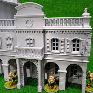 Puede incluir: Un edificio en miniatura gris con puertas arqueadas, balcones y ventanas con contraventanas. Peque&ntilde;as figuras de soldados con uniformes verdes y beige est&aacute;n colocadas alrededor del edificio, lo que sugiere un juego de guerra o un diorama hist&oacute;rico.