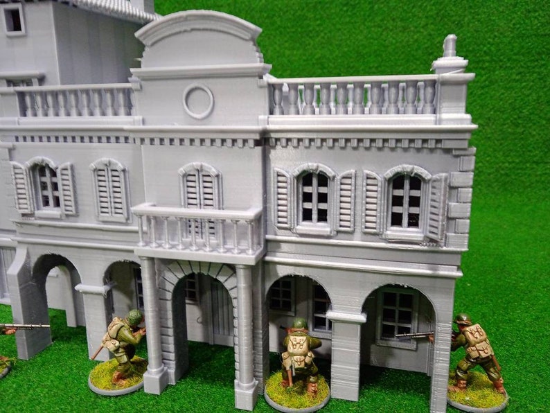 Puede incluir: Un edificio en miniatura gris con puertas arqueadas, balcones y ventanas con contraventanas. Peque&ntilde;as figuras de soldados con uniformes verdes y beige est&aacute;n colocadas alrededor del edificio, lo que sugiere un juego de guerra o un diorama hist&oacute;rico.