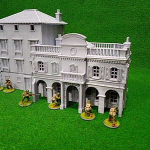 Puede incluir: Un edificio en miniatura gris de varios pisos con detalles arquitect&oacute;nicos, incluyendo ventanas, persianas y un balc&oacute;n. Peque&ntilde;as figuras de soldados est&aacute;n posicionadas alrededor del edificio sobre una superficie verde, sugiriendo un juego de guerra o un diorama.