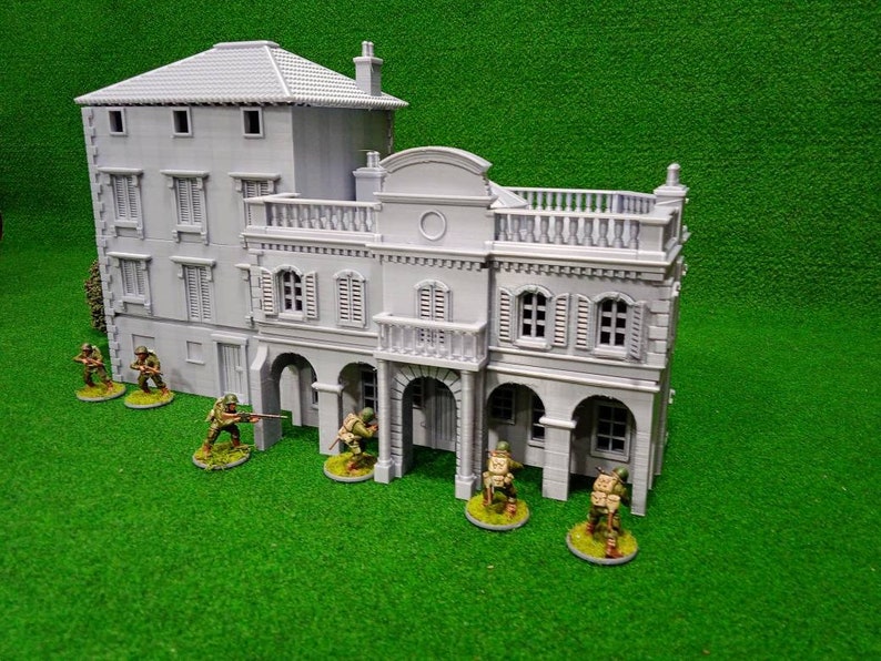 Puede incluir: Un edificio en miniatura gris de varios pisos con detalles arquitect&oacute;nicos, incluyendo ventanas, persianas y un balc&oacute;n. Peque&ntilde;as figuras de soldados est&aacute;n posicionadas alrededor del edificio sobre una superficie verde, sugiriendo un juego de guerra o un diorama.
