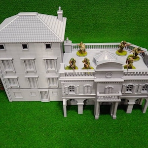May include: A grey, 3D-printed model of a multi-storey building with a tiled roof and multiple windows. Miniature soldiers are positioned on the rooftop. The model is set on a green surface, suggesting a tabletop game or diorama.