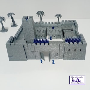 May include: A gray miniature castle model with a tall tower, battlements, and an open gate. Small blue figures are positioned around the castle walls. Palm tree models are placed around the castle. The model is on a white surface.