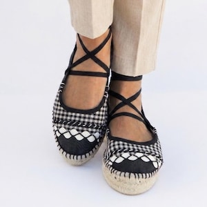 Puede incluir: Bailarinas espadrillas a cuadros blancos y negros con cintas negras cruzadas en el tobillo. Los zapatos tienen una suela de yute tejida y un ribete blanco decorativo en el borde superior. Llevadas con pantalones beige.