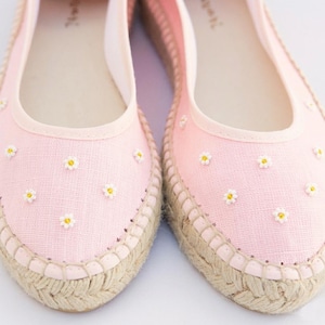 以下が含まれることがあります： 白いデイジー刺繍が施されたピンクのエスパドリーユフラットのペア。靴は、織り込まれたジュートソールと明るいピンクのキャンバスアッパーを備えています。デイジーは黄色の中心を持っています。軽くて夏らしいスタイル。