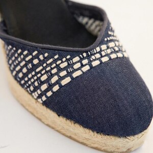 Op de afbeelding: Close-up van een denim espadrille schoen. De schoen heeft een geweven ontwerp in wit en marineblauw over de bovenkant, met een natuurlijke jute zool. De schoen heeft een donker marineblauwe binnenkant.