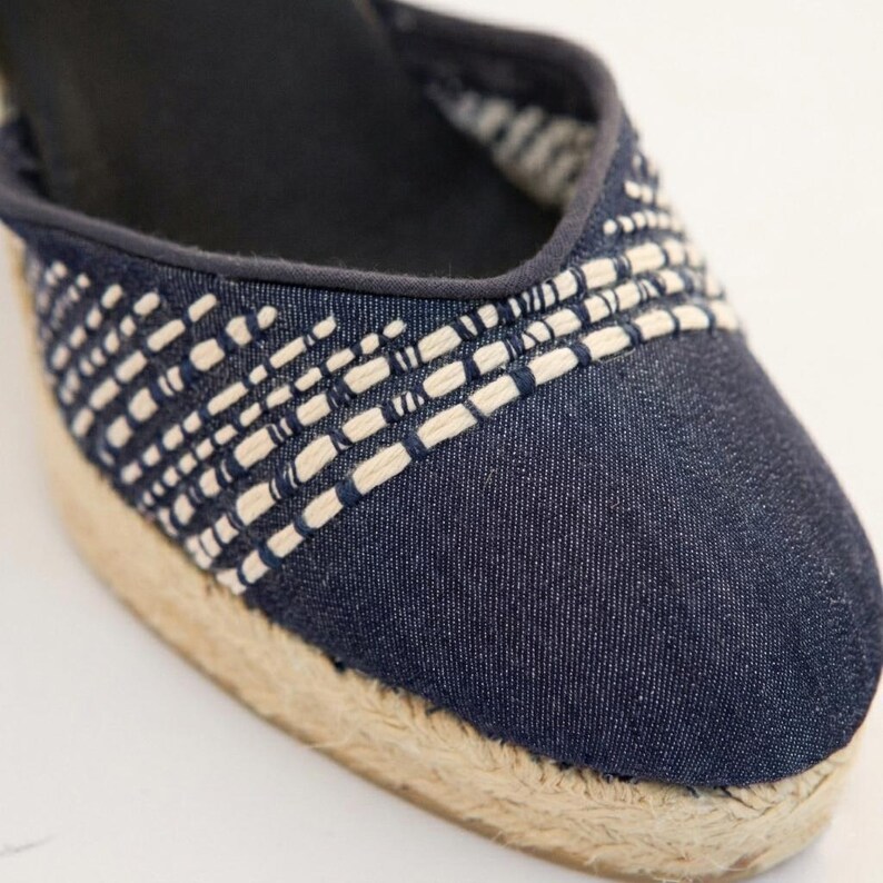 Op de afbeelding: Close-up van een denim espadrille schoen. De schoen heeft een geweven ontwerp in wit en marineblauw over de bovenkant, met een natuurlijke jute zool. De schoen heeft een donker marineblauwe binnenkant.