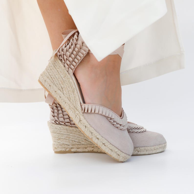 May include: Beige espadrille wedge sandals with woven jute soles and a suede upper. The sandals feature a braided trim and ankle ties. The wedge heel is approximately 3 inches high.