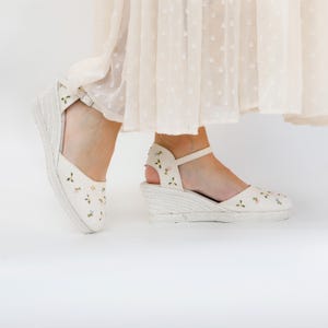 May include: White wedge sandals with a floral embroidery design. The sandals have a closed toe and an ankle strap.