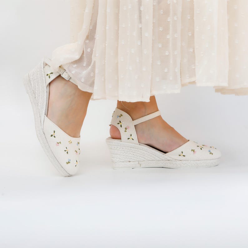 May include: White wedge sandals with a floral embroidery design. The sandals have a closed toe and an ankle strap.