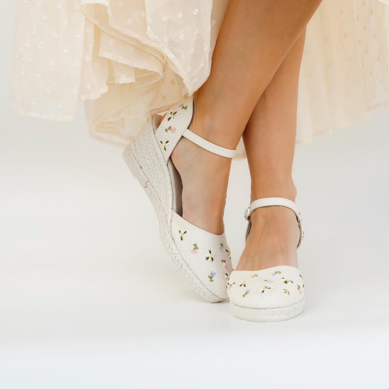 May include: White wedge sandals with a floral embroidery design. The sandals have an ankle strap and a platform sole.