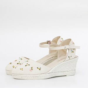 May include: White wedge sandals with a floral embroidered design. The sandals have an ankle strap with a buckle closure.