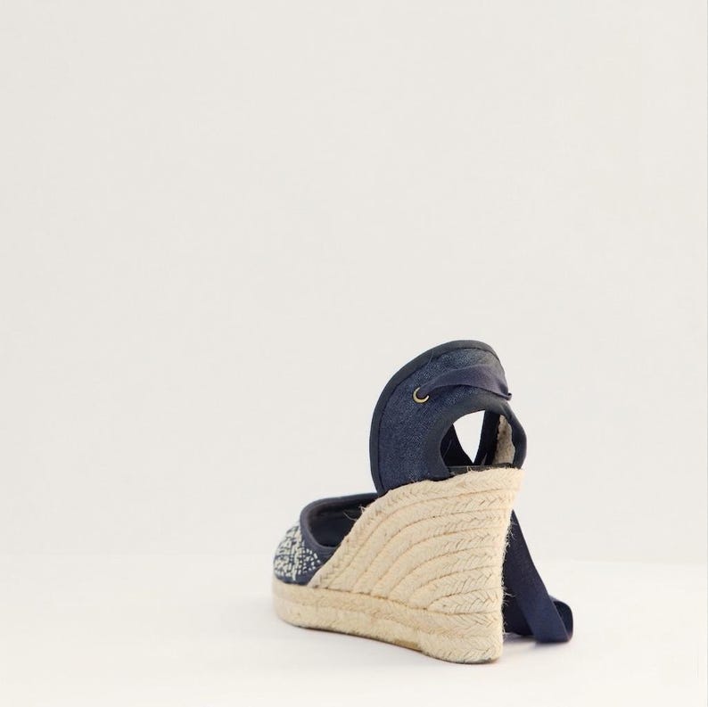 Op de afbeelding: Een spijkerblauwe espadrille sandaal met een geweven jute zool. De sandaal heeft een wikkelenkelband en een decoratief patroon op de teen. De sleehak meet ongeveer 7.6 cm.