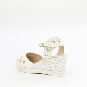 May include: White canvas wedge sandals with a floral embroidery design. The sandals have an ankle strap with a buckle closure.