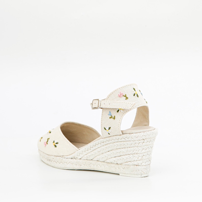 May include: White canvas wedge sandals with a floral embroidery design. The sandals have an ankle strap with a buckle closure.