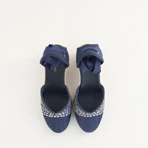Op de afbeelding: Een paar marineblauwe espadrille sleehakken. De schoenen hebben een geweven wit en marineblauw patroon op de neus, een jute zool en marineblauwe enkelbanden van lint. De hakken zijn ongeveer 7.6 cm hoog.