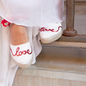 May include: White canvas espadrille shoes with red sequined "love" lettering. The shoes are worn by a person wearing a white dress.