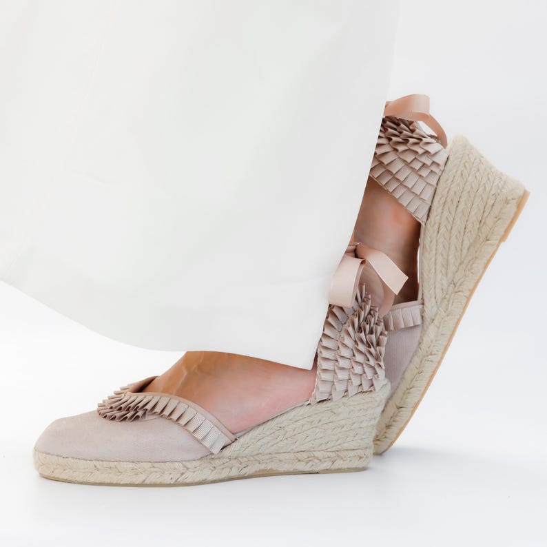 May include: Beige espadrille wedge sandals with a woven jute sole. The sandals feature a ruffled trim and ribbon tie closure. The wedge heel measures approximately 3 inches.