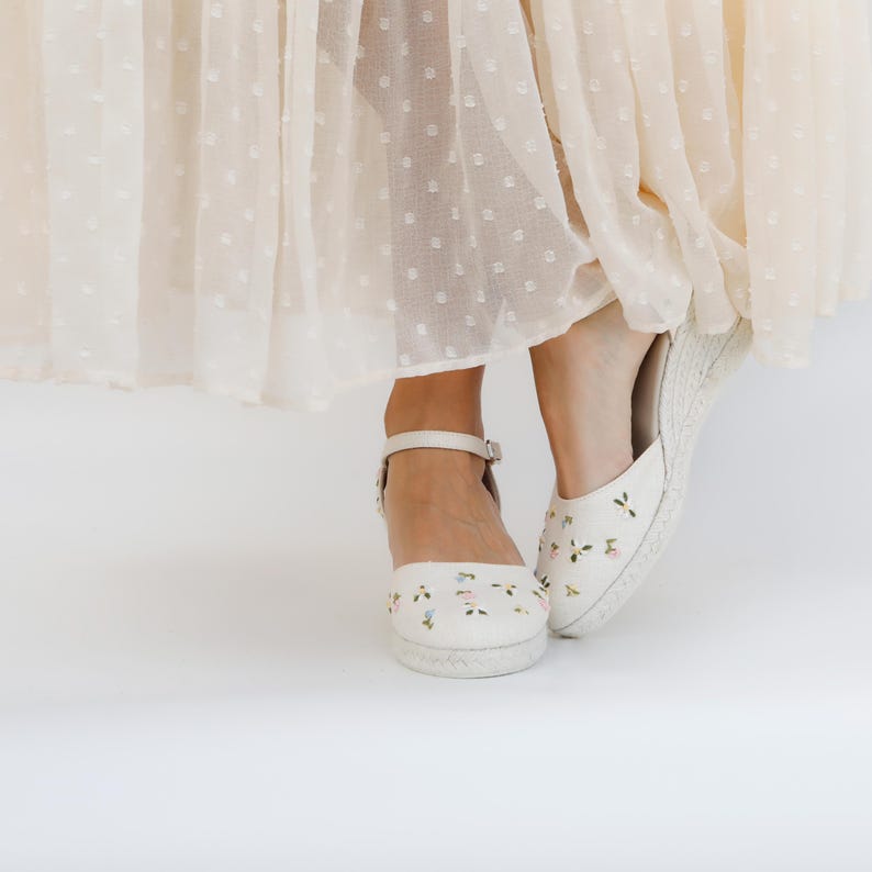May include: A pair of white espadrille shoes with floral embroidery. The shoes have ankle straps and are worn by a person wearing a light beige skirt.