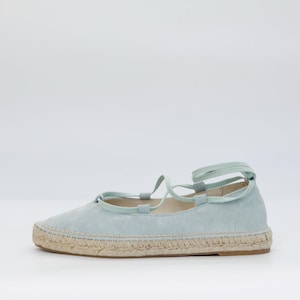 May include: A light blue espadrille flat with a woven jute sole and matching laces. The shoe has a rounded toe and a soft, suede-like upper. The laces wrap around the ankle.