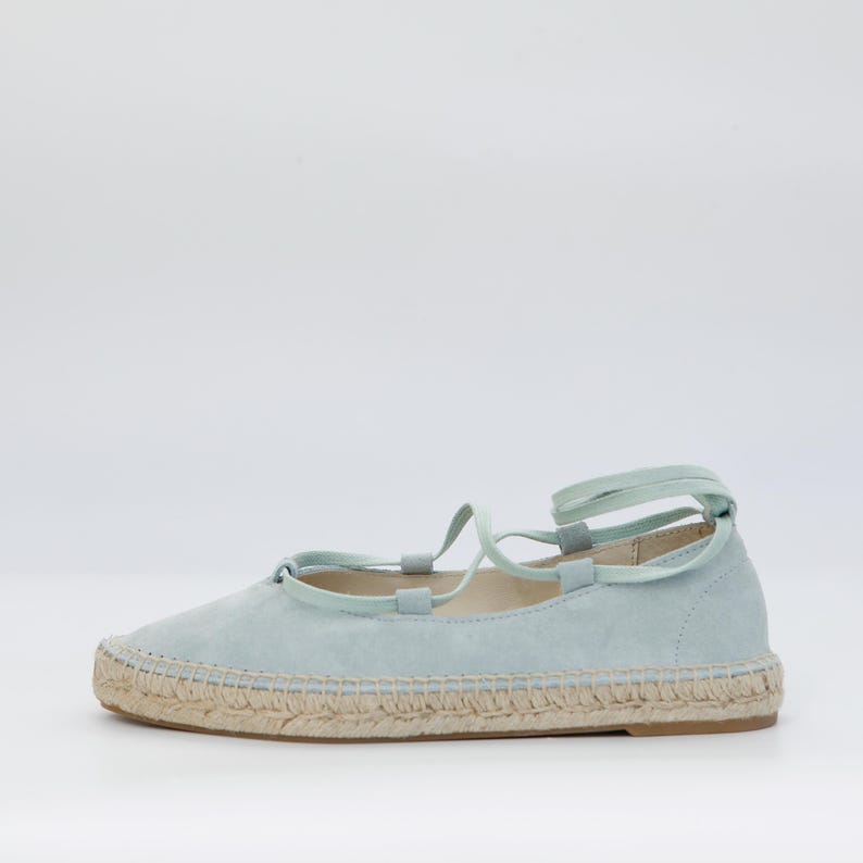 May include: A light blue espadrille flat with a woven jute sole and matching laces. The shoe has a rounded toe and a soft, suede-like upper. The laces wrap around the ankle.
