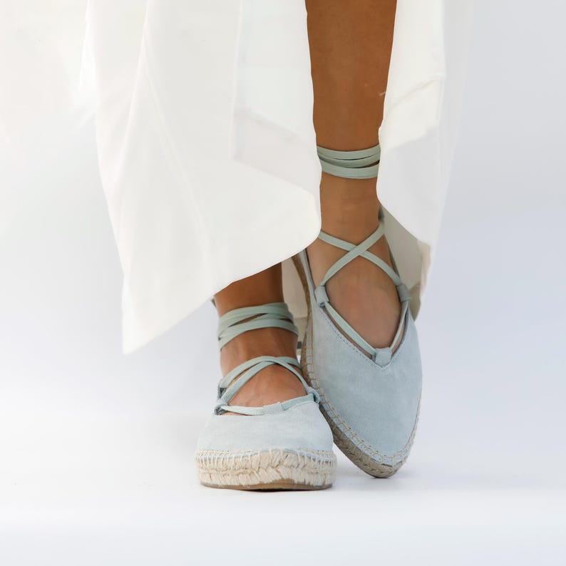 May include: Light blue espadrille flats with matching ankle ties. The shoes have a rounded toe and a woven jute sole. The laces wrap around the ankles. The background is white.