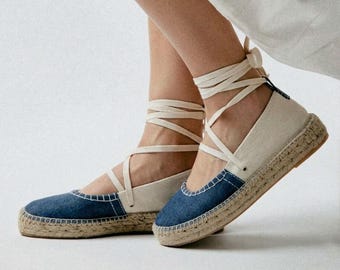 May include: Espadrille flats with denim blue toe caps and natural canvas uppers. The shoes have a woven jute sole and cream-colored wrap-around ankle ties. The shoes are a neutral color and are suitable for casual wear.