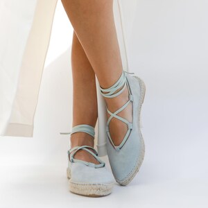 May include: Light blue espadrille flats with ankle ties. The shoes have a suede-like texture and a woven jute sole. The laces wrap around the ankles and cross over the foot. The background is white.