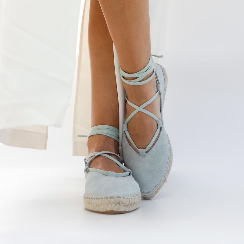 May include: Light blue espadrille sandals with wraparound ankle ties. The sandals have a woven jute sole and a suede upper. The laces are a matching light blue color. The shoes are shown against a white background.