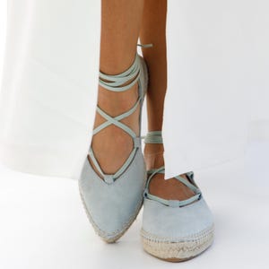 May include: Light blue espadrille flats with a pointed toe and ankle ties. The shoes have a woven jute sole and a soft suede-like upper. The laces are a matching light blue color, creating a cohesive and stylish look.
