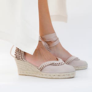 May include: Light pink espadrille wedge sandals with a woven jute sole. The sandals feature a ruffled trim and ankle ties. The wedge heel measures approximately 3 inches.