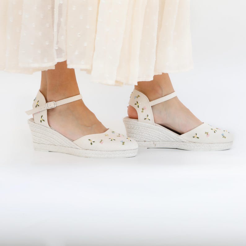 May include: A pair of white wedge sandals with floral embroidery. The sandals have a closed toe and an ankle strap.
