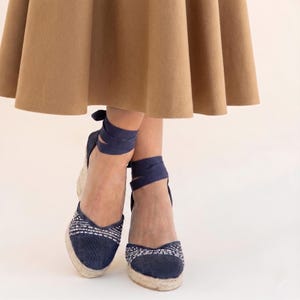 Op de afbeelding: Marineblauwe espadrille sleehakken met geweven jute zolen en wikkelenkelbanden. De neus heeft een wit en blauw geborduurd patroon. Gedragen met een bruine rok.