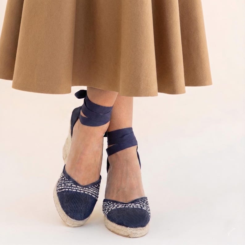 Op de afbeelding: Marineblauwe espadrille sleehakken met geweven jute zolen en wikkelenkelbanden. De neus heeft een wit en blauw geborduurd patroon. Gedragen met een bruine rok.