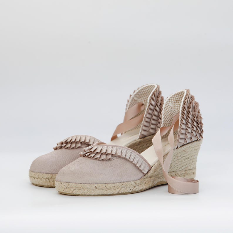 May include: A pair of blush-colored espadrille wedges with a woven jute sole. The shoes feature a suede upper with ruffled trim and ribbon ankle ties. The wedge heel provides a comfortable lift.