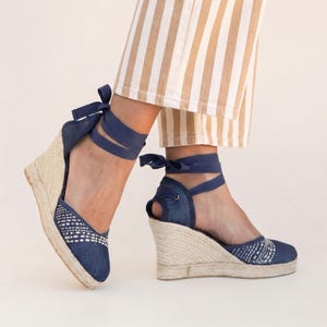 Op de afbeelding: Denim espadrille sleehak sandalen met geweven details en enkelbanden. De sandalen hebben een jute touwzool en een marineblauwe stoffen bovenkant. De enkelbanden zijn marineblauw. De sleehak is ongeveer 7.6 cm hoog.