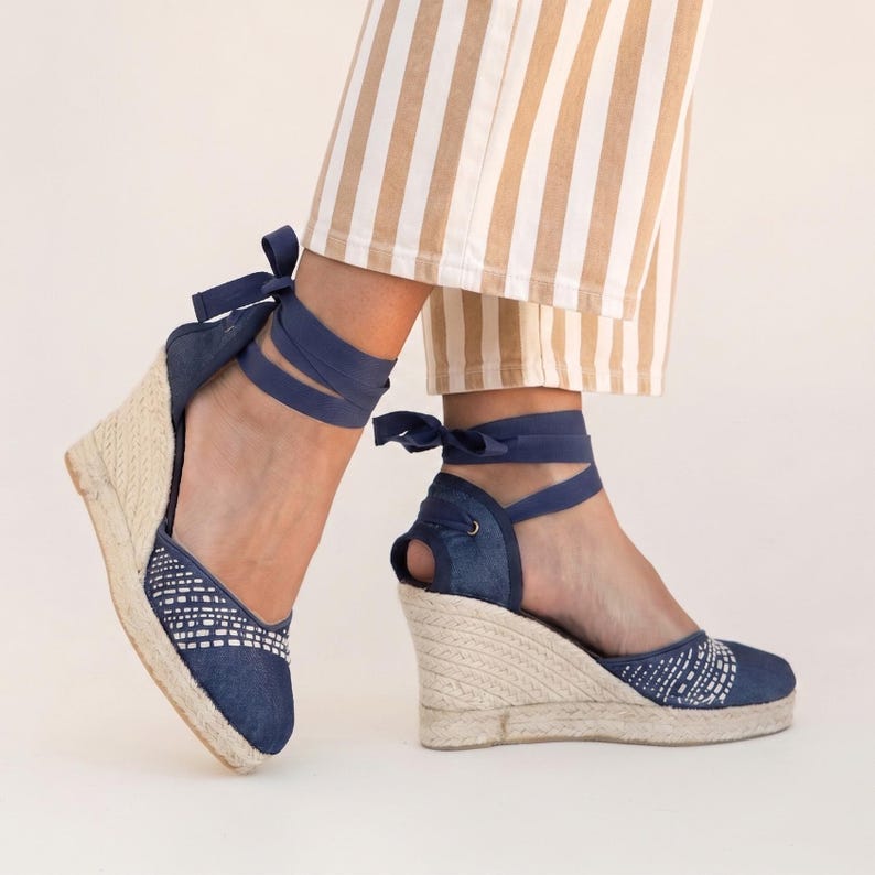Op de afbeelding: Denim espadrille sleehak sandalen met geweven details en enkelbanden. De sandalen hebben een jute touwzool en een marineblauwe stoffen bovenkant. De enkelbanden zijn marineblauw. De sleehak is ongeveer 7.6 cm hoog.