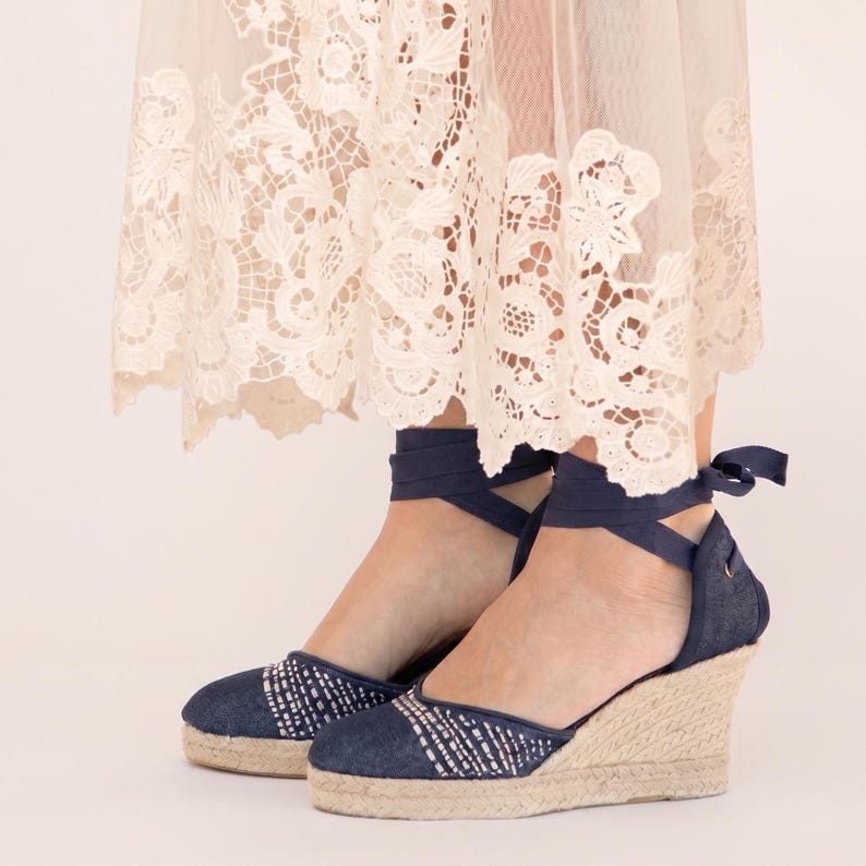 Op de afbeelding: Spijkerstof wig espadrilles met marineblauwe enkelbanden. De schoenen hebben een geweven jute zool en een wit en blauw patroon op de neus. De sleehak biedt hoogte en ondersteuning.