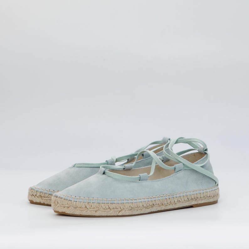 May include: A pair of light blue espadrille flats with woven jute soles and ankle ties. The shoes have a suede-like texture and a minimalist design, suitable for casual wear. The soles are approximately 1 inch thick.