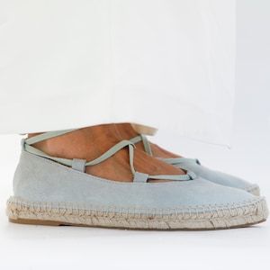 May include: Light blue suede espadrille flats with a woven jute sole and matching laces. The flats have a rounded toe and a low profile, perfect for warm weather. The laces wrap around the ankle.
