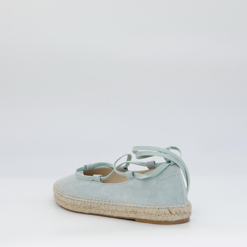 May include: A light blue espadrille flat with a woven jute sole and matching laces. The shoe has a rounded toe and a soft suede-like upper. The laces wrap around the ankle for a secure fit. A casual and stylish footwear option.