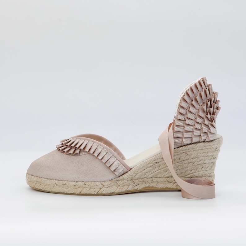 May include: A pair of blush-colored espadrille wedges. The shoes feature a suede upper with a ruffled trim, a woven jute sole, and a ribbon ankle tie. The heel height is approximately 3 inches.